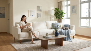 Woman reading on a cream 3-seater sofa in a bright jumbo HDB living room with coffee table, rug, plants, and cat.
