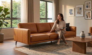 Woman reading on a tan aniline leather sofa in a warm Singapore living room with large windows and indoor plant.