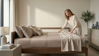 Woman making a bed with neutral-toned bedsheets on a wooden bed frame, demonstrating proper fitted sheet coverage and mattress fit in a modern Singapore bedroom.