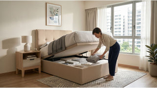 Woman storing bedding inside a beige storage bed in a bright HDB bedroom with large windows