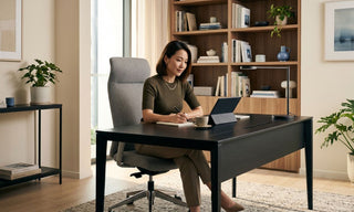 Woman working at a black desk in a warm Singapore home office with bookshelf, plants and grey office chair
