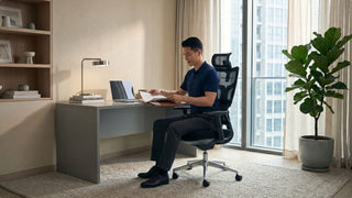Man using a black ergonomic office chair with desk, lamp, books, and warm lighting in a modern Singapore home office
