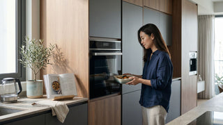 Woman placing a dish into a built-in oven installed in a modern Singapore kitchen with wood cabinetry.
