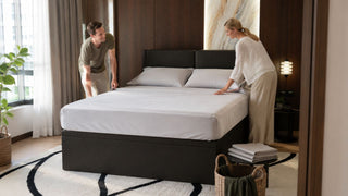 White couple arranging fresh bedsheets on a dark upholstered bed in a modern Singapore bedroom
