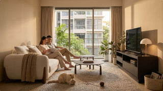 Couple reading on a cream sofa in a warm condo living room with balcony, coffee tables, TV console, plants, and pet cat