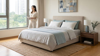 Woman standing beside a cooling mattress in a bright Singapore bedroom with soft blue bedding and neutral decor.