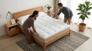 Couple placing a white mattress topper on a light wood bed frame in a bright Singapore bedroom