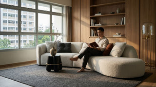Man reading on a cream bouclé sectional sofa in a modern Singapore living room with wood shelves and large windows
