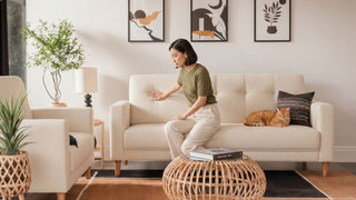 Woman checking a cream fabric sofa cushion in a bright living room with cat, rattan coffee table, plants, and wall art