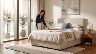 Woman arranging bedding on a beige divan base in a bright Singapore condo bedroom with neutral decor.