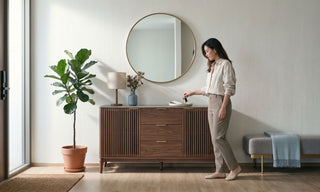 Woman placing keys on a wooden entryway cabinet with round mirror, indoor plant, lamp, and bench in a modern Singapore home.