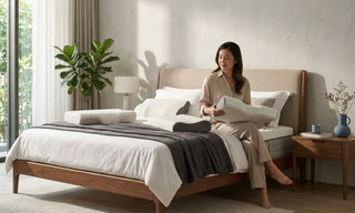 Woman comparing ergonomic pillows on a bed in a Singapore bedroom with natural light and modern wooden furniture.