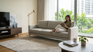 Woman reading on a beige foldable sofa in a bright Singapore condo living room with TV console and balcony view.
