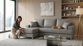 Woman adjusting a grey chaise sofa in a modern condo living room with cat, glass coffee table, shelves, and city skyline view