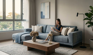 Woman reading on a grey L-shaped sofa in a modern Singapore living room with coffee table and large windows.