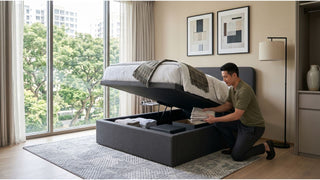 Man storing folded linens inside a grey storage bed with gas lift mechanism in a bright bedroom