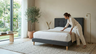 Woman styling a grey upholstered king bed frame in a landed home bedroom with garden views, neutral bedding, and soft natural light
