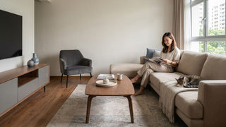 Woman reading on a beige sofa in a warm wood HDB living room with coffee table, TV console, and cat.
