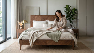 Woman styling a herringbone wood bed frame in a calm Singapore bedroom with neutral bedding and warm timber furniture.