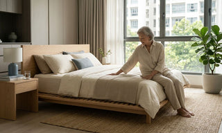 Elderly woman sitting on a low Japanese tatami mattress in a bright Singapore bedroom with a slatted wooden base.