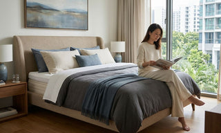 Woman reading on a beige upholstered bed with layered pillows in a bright Singapore condo bedroom.