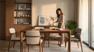 Woman styling a warm Liang Wood dining room with wooden table, upholstered chairs, built-in shelves, and soft natural light.