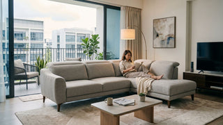 Light grey L-shape sofa in a Singapore condo living room with balcony view, coffee table, TV console and woman reading on the chaise.
