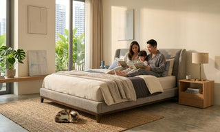 Family reading together on a low platform bed in a warm Singapore condo bedroom with neutral bedding, wood side table, indoor plants, and a pet cat.