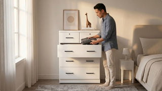 Man placing folded clothes into a white drawer cabinet in a warm modern bedroom