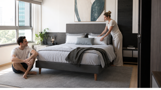 Modern grey bed frame in a bright Singapore condo bedroom with a couple arranging cushions for a practical home setup