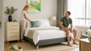 Modern Singapore bedroom with a grey upholstered divan bed frame, white bedding, and a couple arranging cushions in a bright HDB-style room.