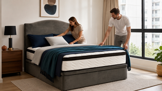 White couple arranging bedding on a grey upholstered luxury bed frame in a modern Singapore condo bedroom.