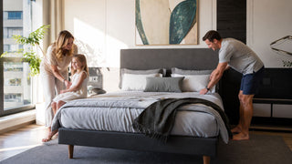 White family arranging a queen mattress in a modern Singapore bedroom with a charcoal upholstered bed frame and soft grey bedding.