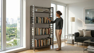 Woman arranging books on a metal bookcase beside large windows in a modern condo