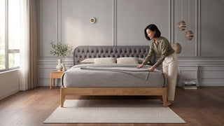Woman arranging bedding on a minimalist wooden bed frame with grey upholstered headboard