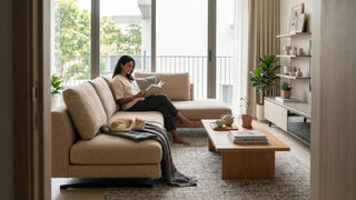 Minimalist Singapore condo living room with beige sofa, oak coffee table, TV console, balcony view, and a woman reading with a cat