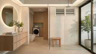 Modern HDB toilet renovation with wood vanity, round mirror, glass shower screen, indoor plants, and warm neutral tiles.