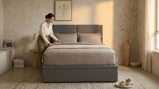 Woman arranging a neutral queen size bedsheet on a grey upholstered bed in a cosy Singapore bedroom with soft daylight