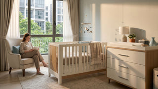 Mother reading beside a baby cot, changing dresser and nursing chair in a calm Singapore nursery bedroom.