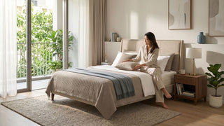 Woman arranging pillows on a pocketed spring mattress in a bright Singapore condo bedroom with neutral bedding and balcony greenery.