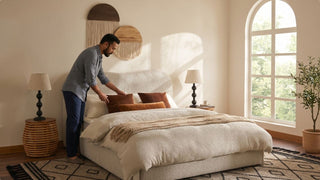 Man arranging pillows on a queen bed frame in a cosy condo bedroom with woven bedside table