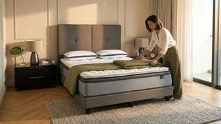 Woman folding a blanket on a queen mattress in a cosy Singapore bedroom with grey upholstered bed, pillows, and balcony light