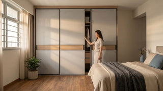 Woman opening a sliding-door wardrobe in a compact 3-room HDB bedroom with warm wood flooring and neutral bedding.
