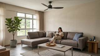 Woman reading on a grey sectional sofa in a Singapore HDB living room with a black ceiling fan, coffee table and indoor plant.