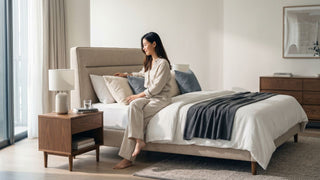 Woman styling cushions on a soft modern bed with upholstered headboard, wooden bedside table, and neutral bedroom decor in Singapore.