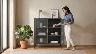 Woman arranging decor in a black steel storage cabinet with glass doors in a bright Singapore living room.