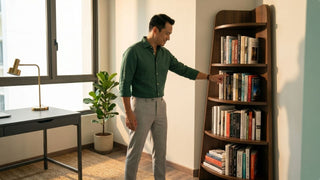 Man browsing a tall dark wood corner bookshelf in a warm modern study with a desk and indoor plant
