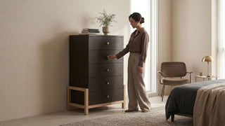Woman checking a tall dark wood storage drawer unit in a neutral bedroom with soft lighting and modern furniture