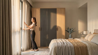 Woman adjusting sheer curtains beside a wooden wardrobe to protect it from afternoon sun fade in a Singapore bedroom.