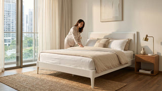 Woman arranging neutral bedding on a white bed frame in a bright Singapore condo bedroom.